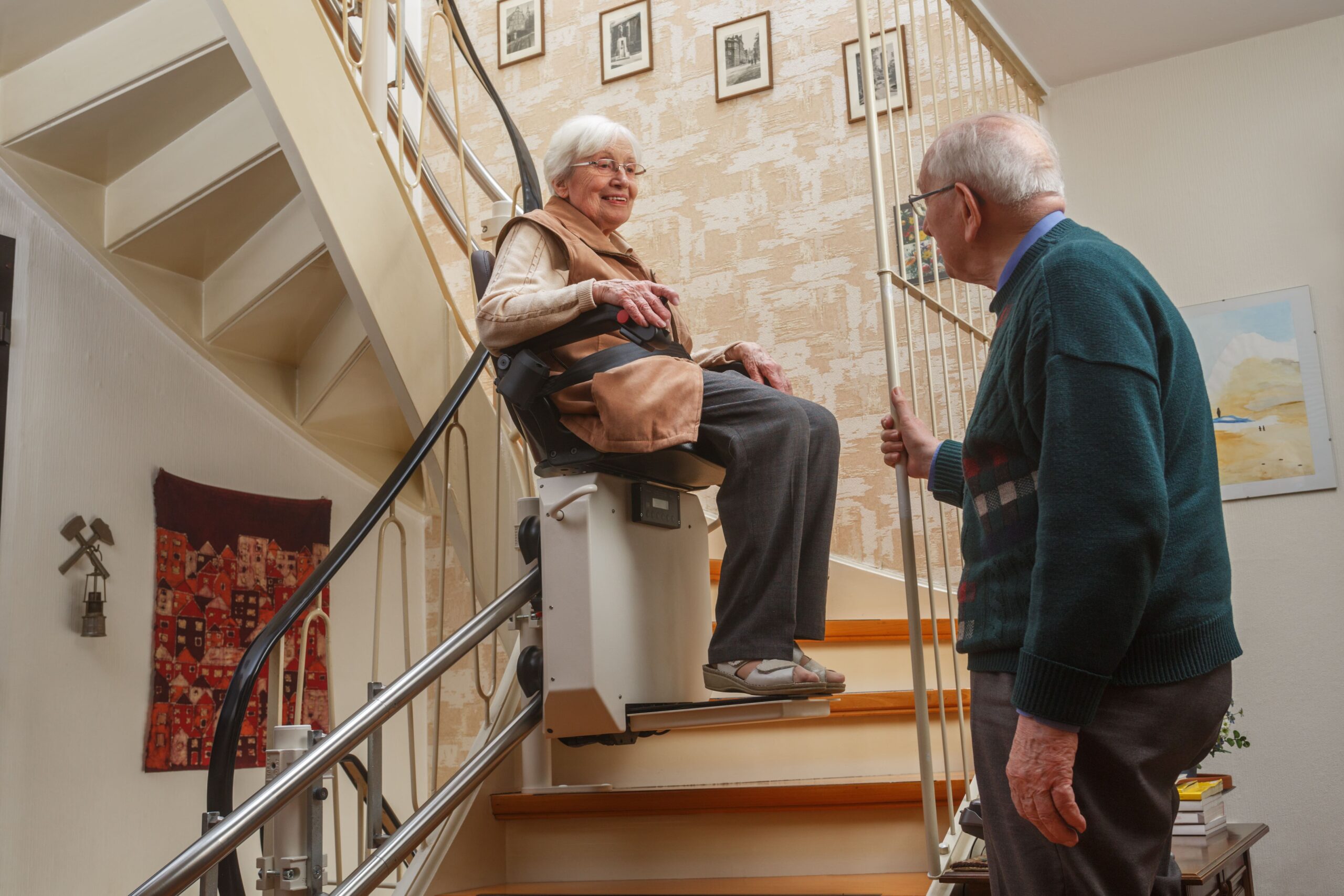 older woman sitting in a Stair Lift while talking to an older man who is standing at the bottom of the stairs